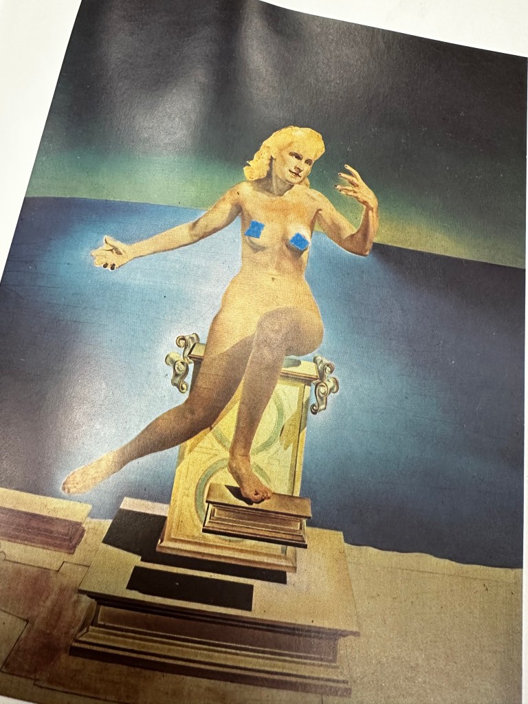 A surreal painting depicts a nude figure with blonde hair sitting atop a stack of books against a gradient blue-green background, arms outstretched expressively. The image, a photograph of a book page, features blue tape over each breast of the figure.