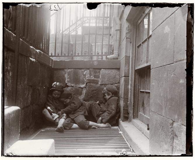 Three young boys in worn clothing and no shoes sit on a metal grate in a narrow, stone-walled alley, with barred windows and steps visible in the background.