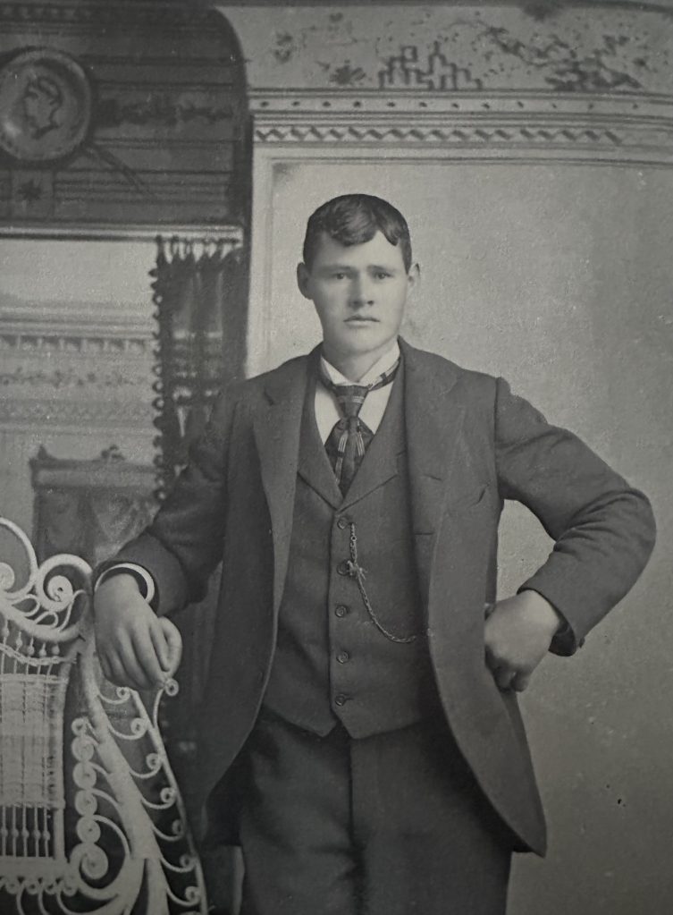 A vintage black-and-white photograph of a young man posing indoors in a formal three-piece suit. He stands confidently with one hand on his hip and the other resting on an ornate, white wicker chair. He wears a dark jacket, a buttoned vest with a pocket watch chain, a light collared shirt, and a patterned necktie against a decorated room backdrop.