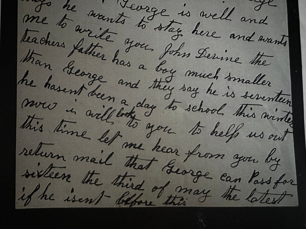 A close-up photograph of a handwritten letter in dark ink on light-colored paper. The elegant, cursive handwriting features long, sweeping loops and slanted lettering characteristic of older historical correspondence.The visible portion of the letter reads: George is well and he wants to stay here and wants me to write you John Devine the teachers father has a boy much smaller than George and they say he is seventeen he hasent [sic] been a day to school this winter now I will look to you to help us out this time let me hear from you by return mail that George can pass for sixteen the third of May the latest..."